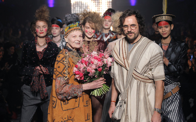 Dame Vivienne Westwood and Andrew Kronthaler posing with flowers following their fashion show to raise awareness for indigenous rights. Fashion models are in the background.