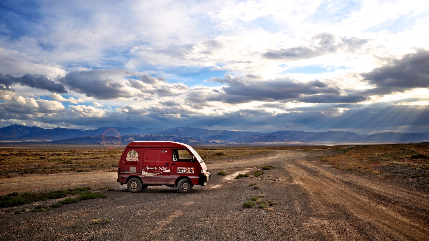 A very tired looking bedford rascal van is parked in a vast empty plain under a brooding sky.