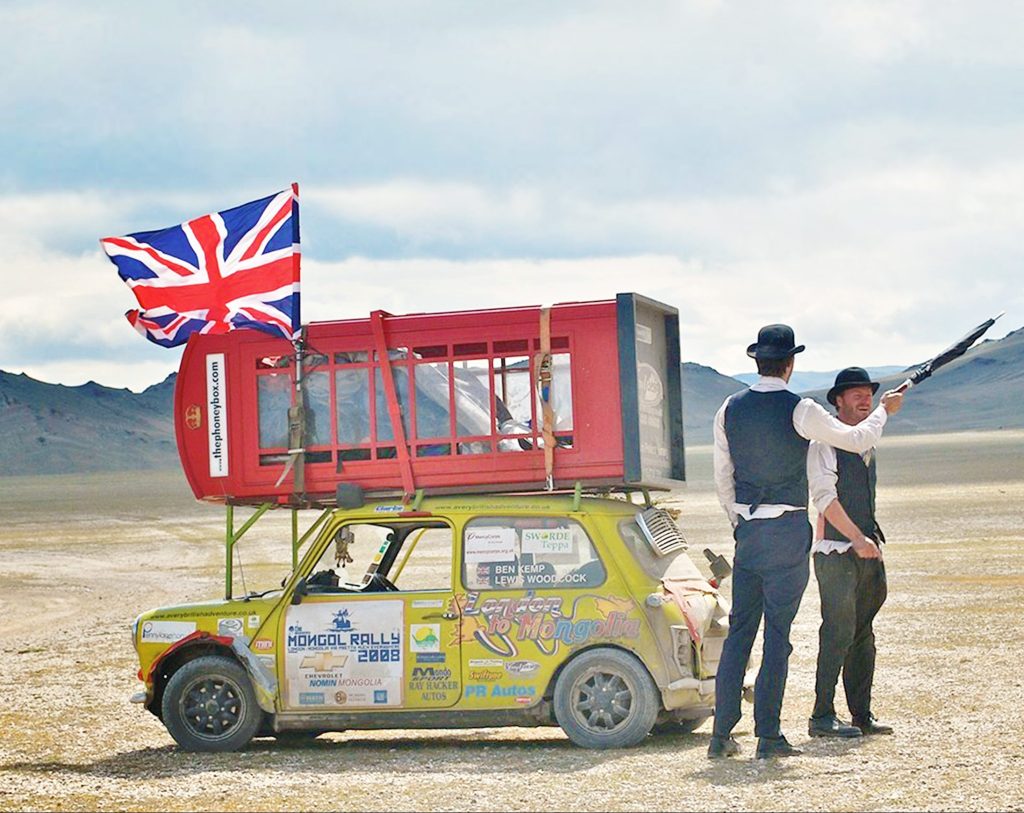 Une cabine téléphonique rouge est attachée au toit d'une mini cooper jaune, garée dans la steppe mongole. À proximité, deux messieurs en chapeau melon font un geste vers une montagne avec un parapluie. Telle est la vie sur le rallye mongol.