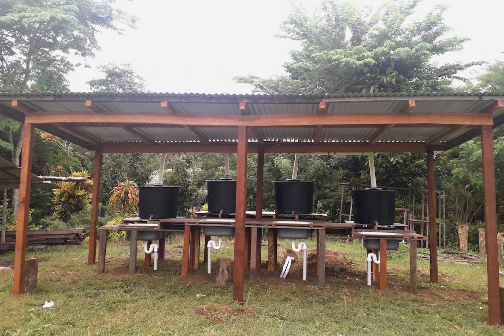 Washing hands fights deforestation. Four handwashing stations sit on tables under a corrugated roof, in a small clearing of rainforest. 