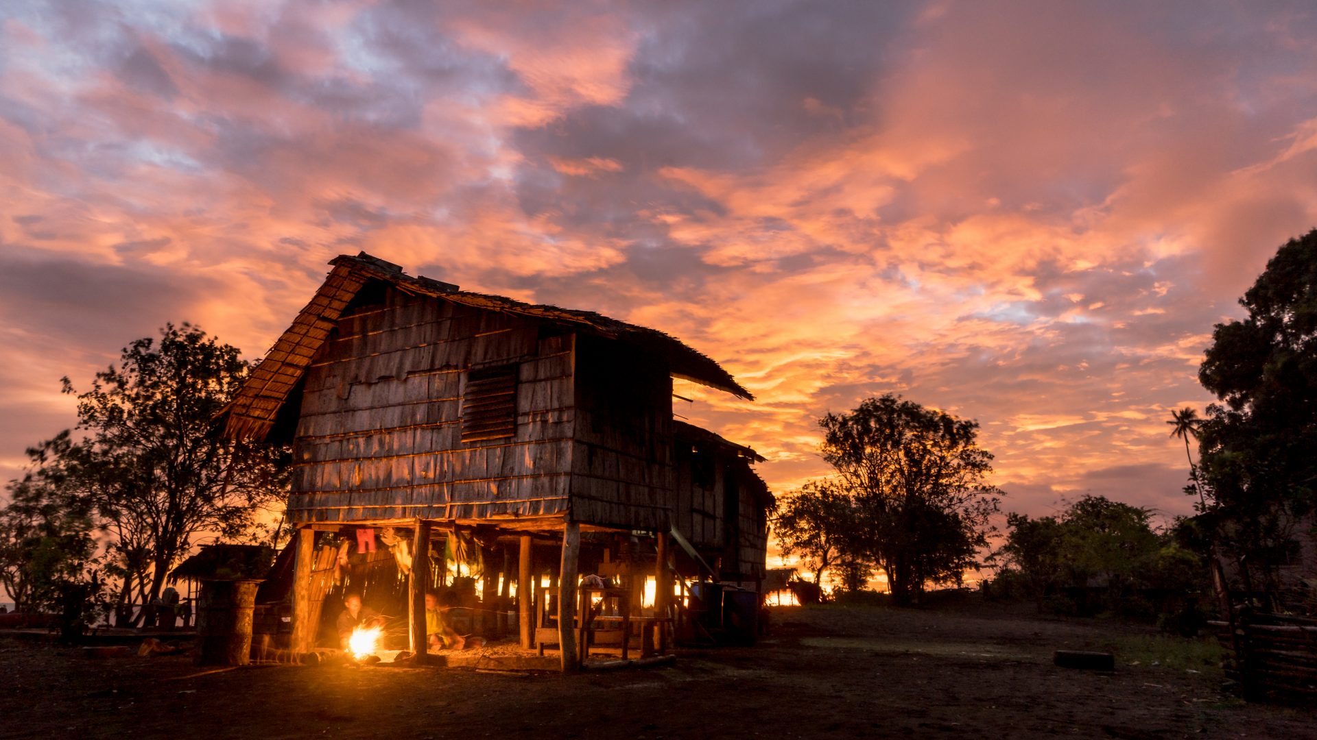 Une maison côtière recouverte de bois sur pilotis se trouve derrière un magnifique coucher de soleil rouge et orange. Il y a des arbres qui se profilent et vous pouvez juste distinguer un petit feu de cuisine allumé avec deux personnes rassemblées autour de lui. L'image semble paisible.