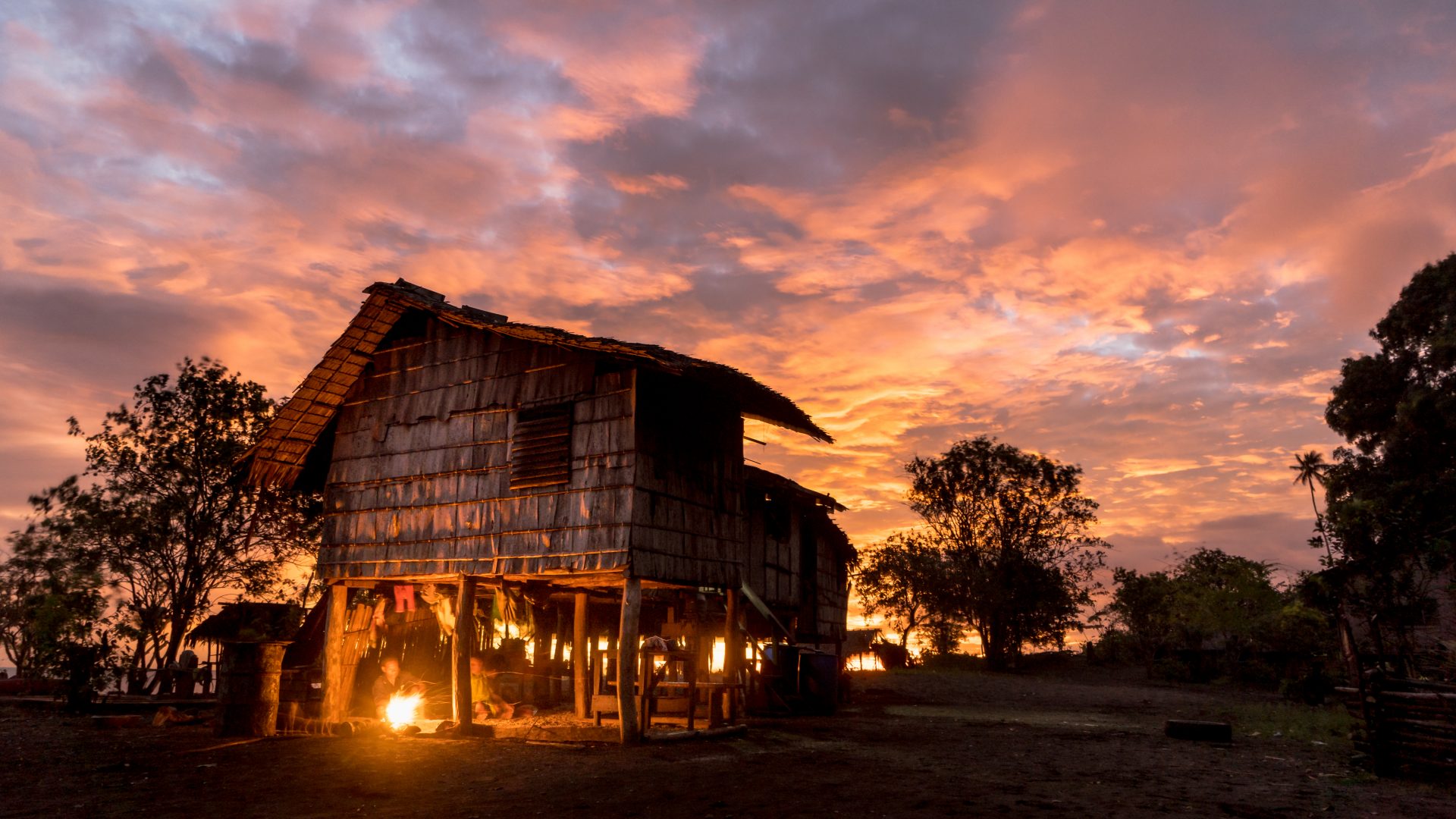 A wood cladded, coastal house on stilts sits has a beautiful red and orange sunset behind it. There are trees that are silhouetted and you can just make out a small cooking fire burning with two people gathered around it. The image feels peaceful.