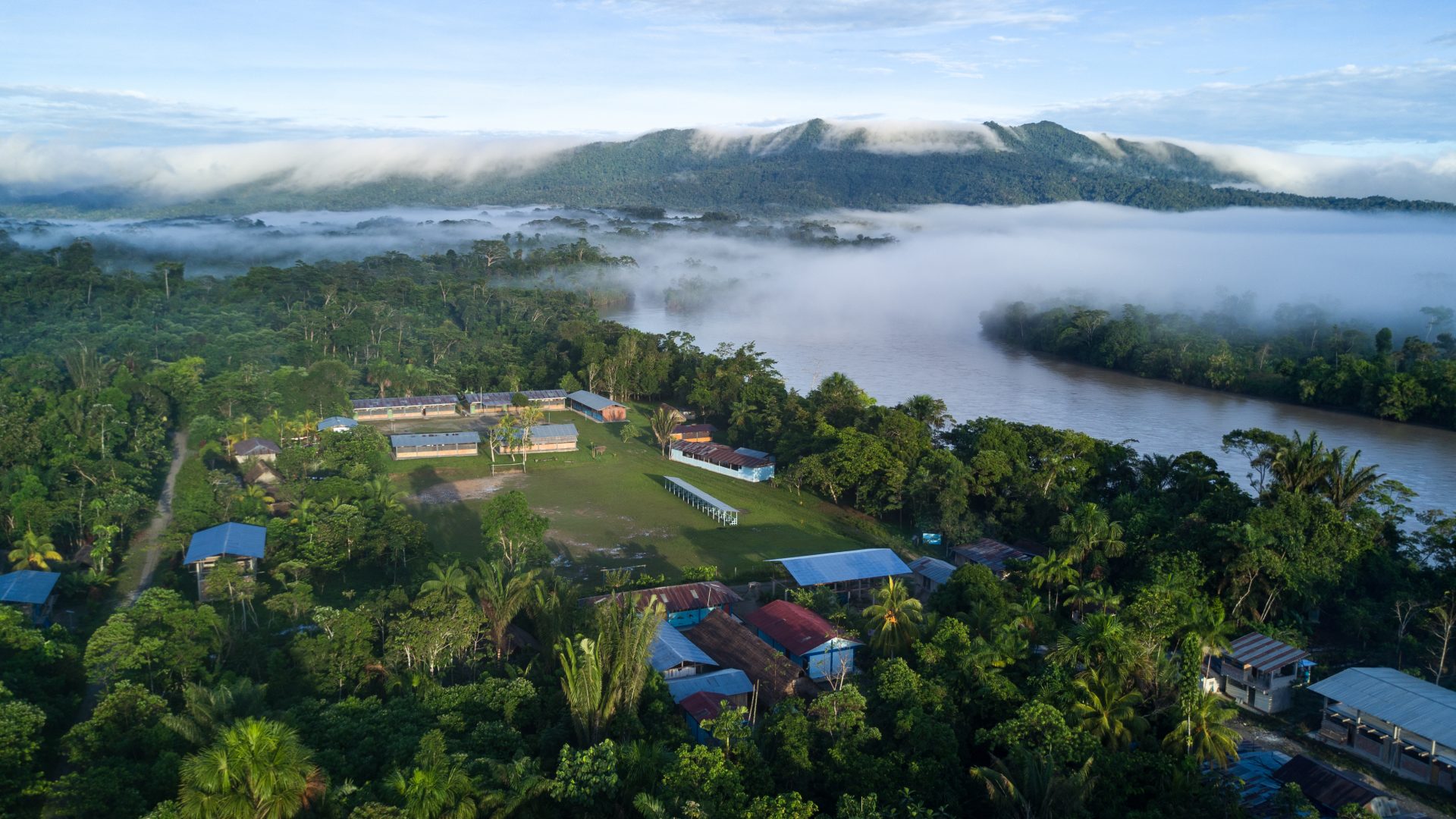 Aerial image of a village near a river with forested mountains in the background.