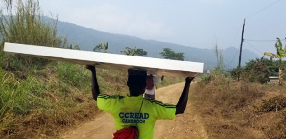 A CCREAD staff member balancing a solar panel on their head, walking down a road with misty hills and the Congo Rainforest in the background.