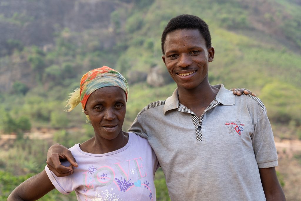 A mother and son are pictured in front of the rainforest, with their arms around each other, facing the camera and smiling.