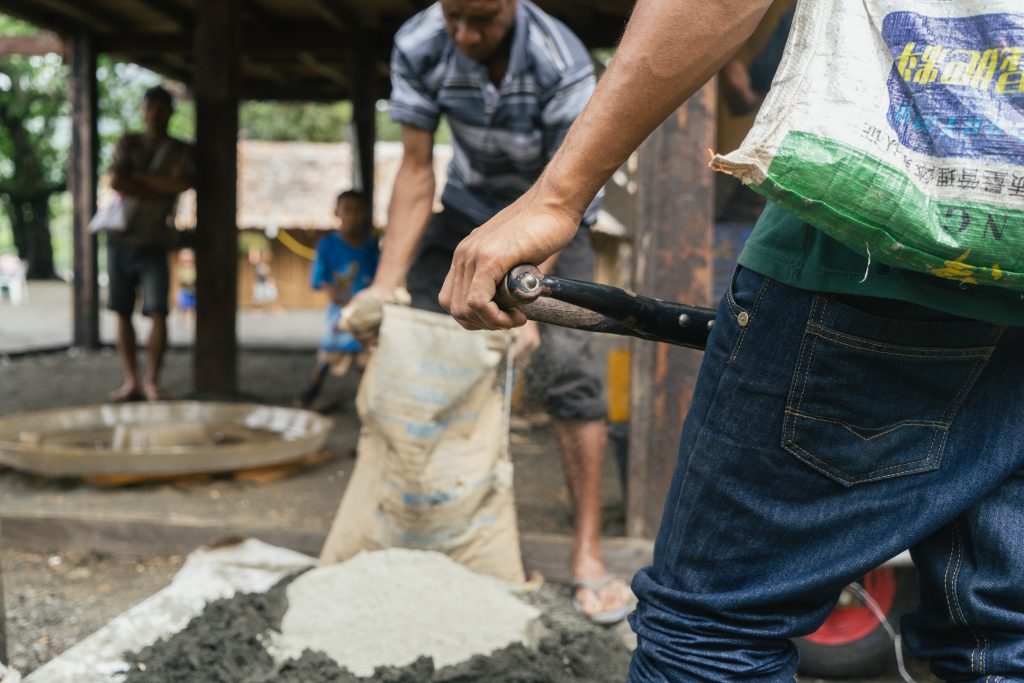 Closeup of a hand holding the handle of a shovel whilst a bag of cement is emptied in the background.