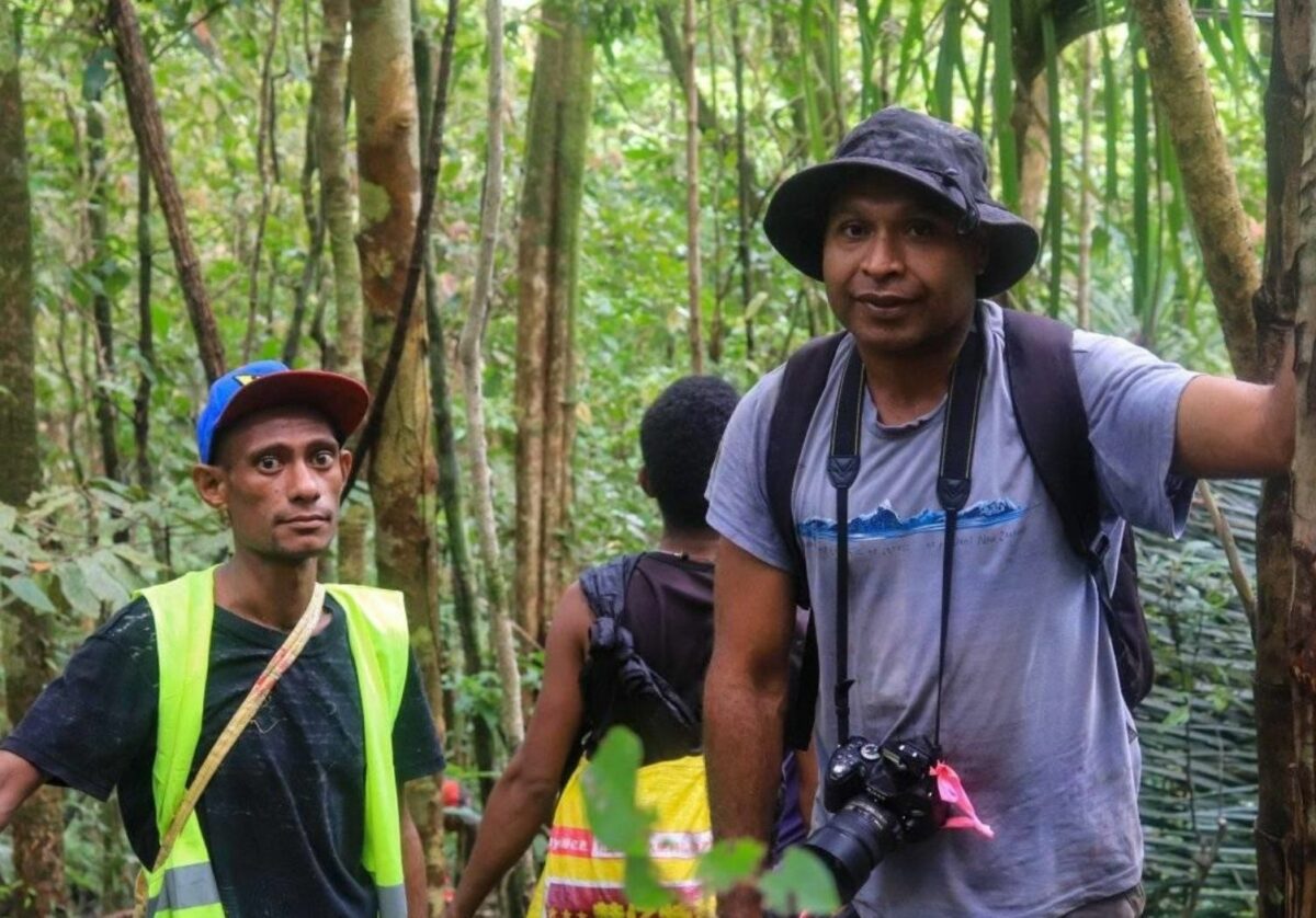 Standing in a forest is Dr Chris Dahl on the right wears a blue tshirt with a balck backpack, black hat and a camera around his neck, next to him his biodiversity officer Jack Joseph Kostolo, he wears a blue cap, black tshirt and a high vis jacket