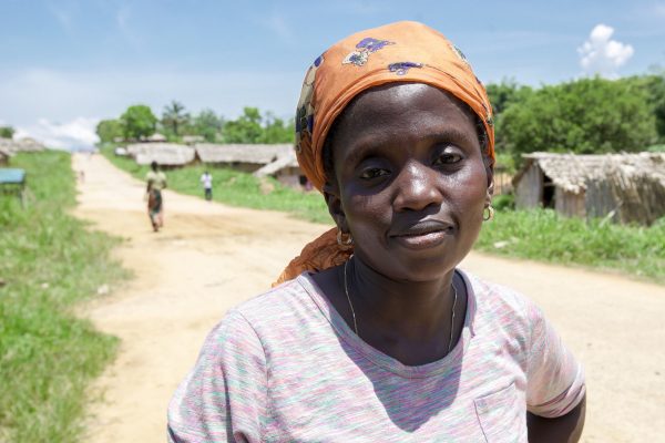 Lady from Lubutu community with orange head scarf and purple t-shirt looks at the camera, she is standing on a dirt road, with green verges, behind her are thatched houses and forest in the distance