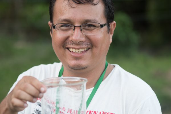 A man (Josue) smiles at the camera whilst holding a glass beaker