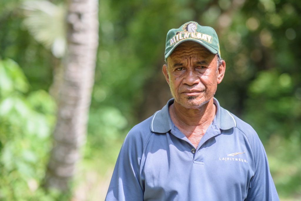 Community member portrait he is wearing a blue shirt and green hat, with forest in the background 