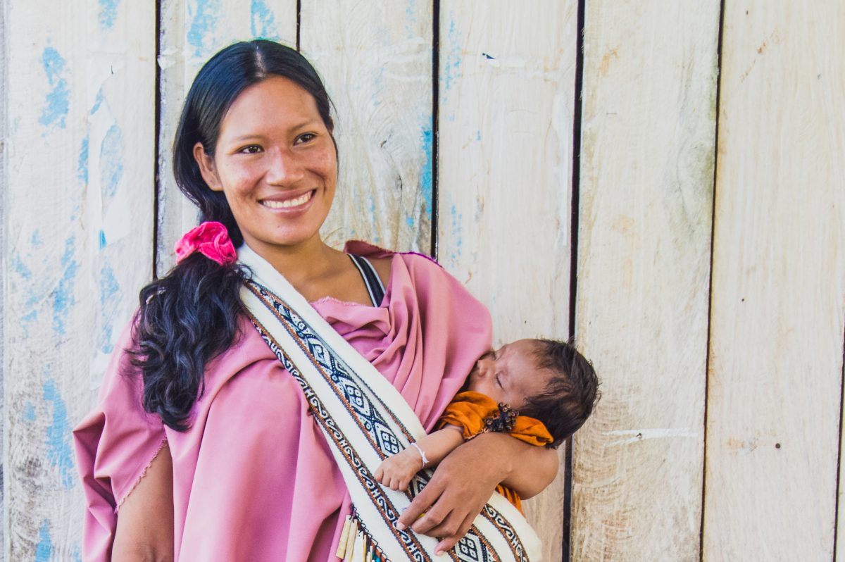 Portrait of Adelaida, She is smiling and wears a pink dress, with her hair pulled into a side ponytail with matching pink hair scrunchy , she holds a baby in her arms