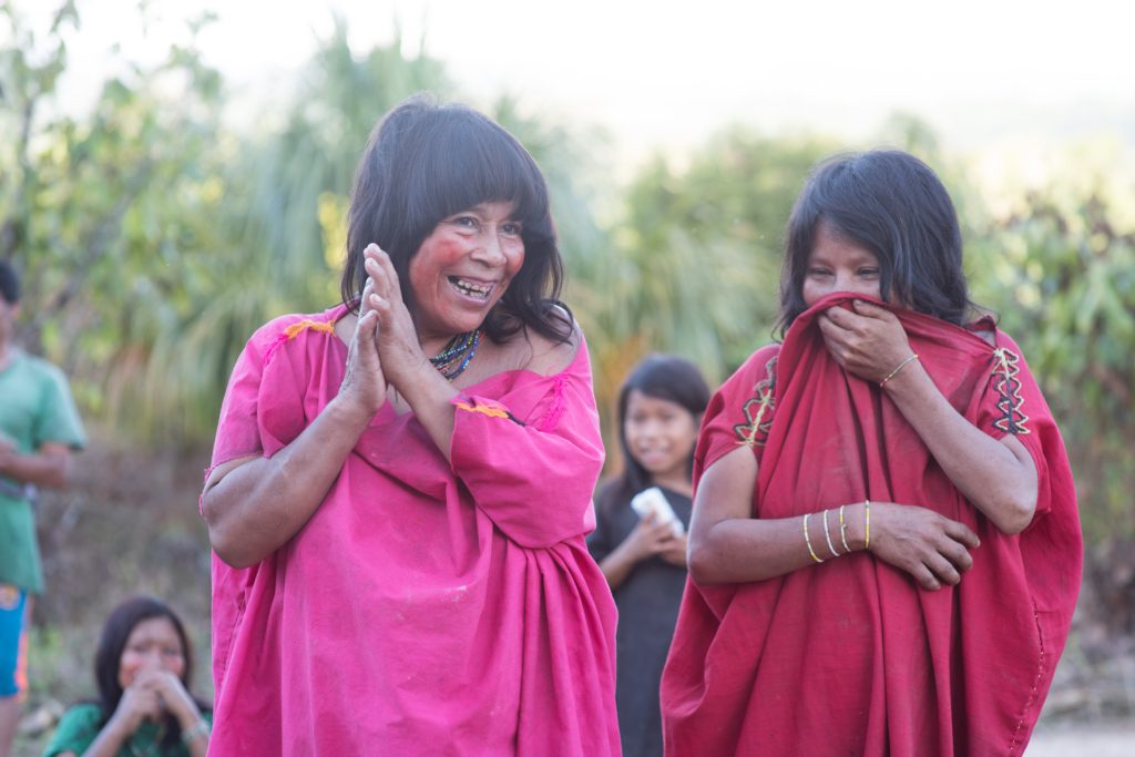 Two women from the Asháninka community, they wear pink dress, one lady is smiling while the other lady covers her mouth. It is a bright scene.
