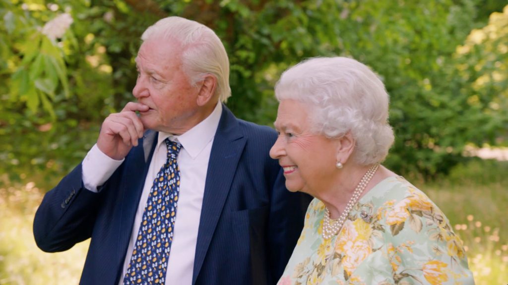 Sir David Attenborough wearing a suit and tie standing with HM the queen wearing a mint and yellow floral dress. Surrounded by trees. in the gardens of Buckingham Palace.