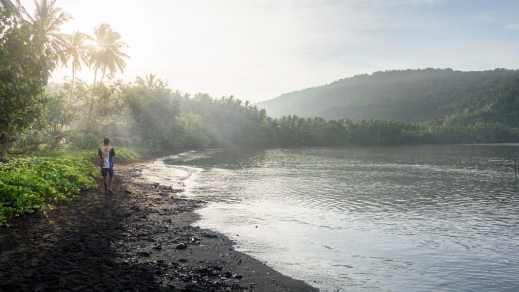 A man walks down the river bank in PNG. Rainforest lines the river and sunlight filters though.