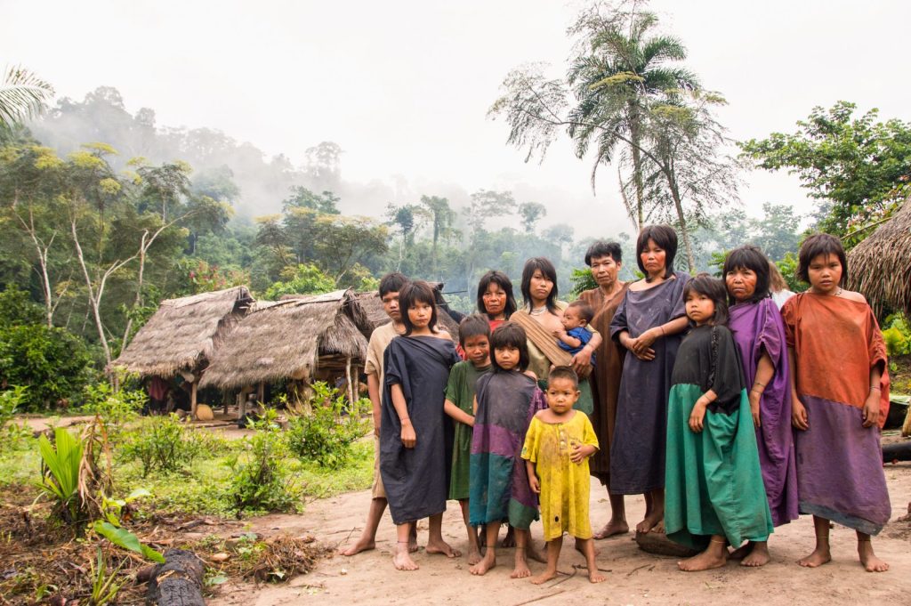 Members of the Ashaninka community including adults and children stand together in front of houses, a mist hangs in the air and rainforest can be seen in the background