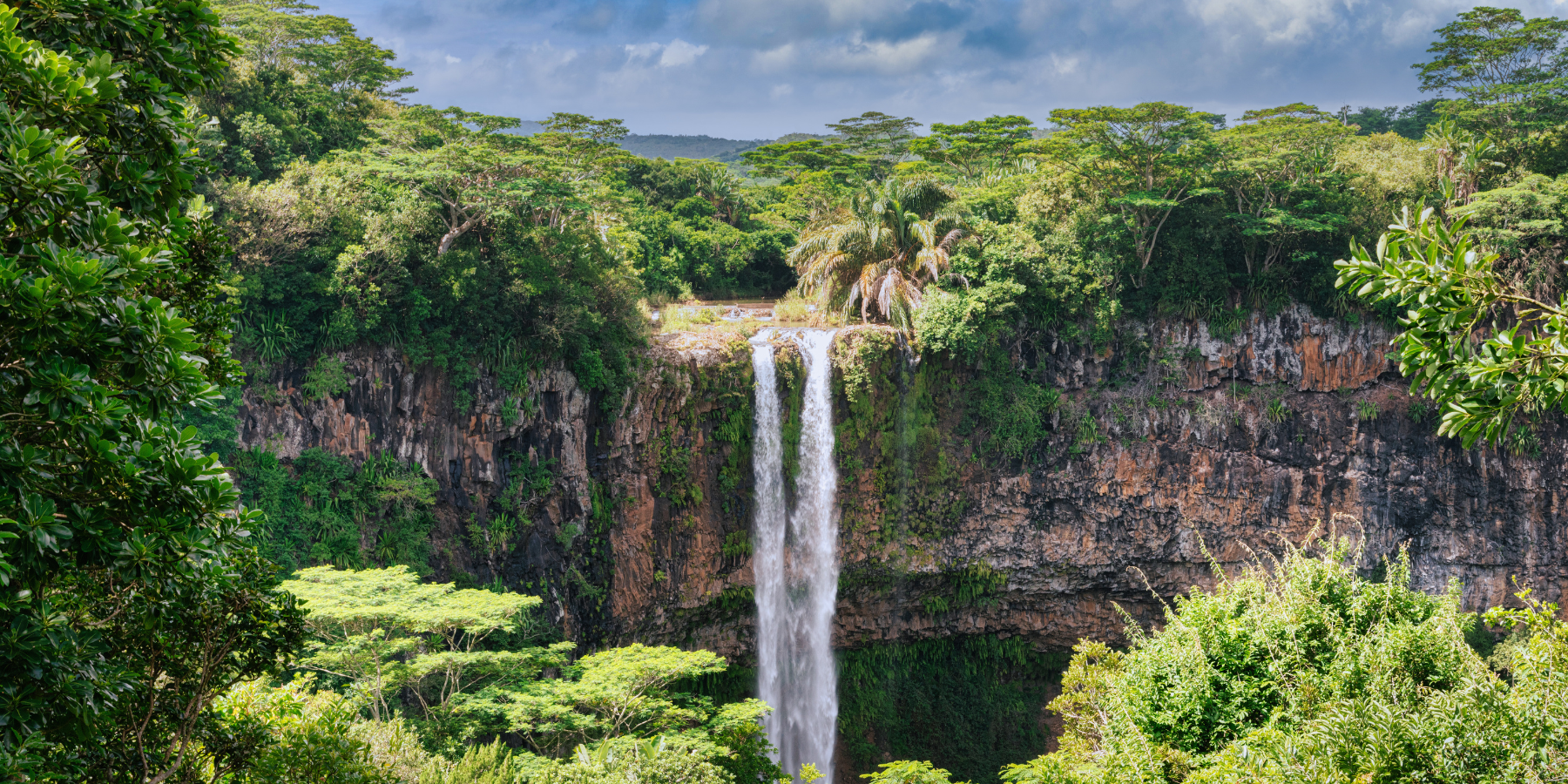 A waterfall spills over a cliff edge, surrounded by lush rainforest with a blue sky above