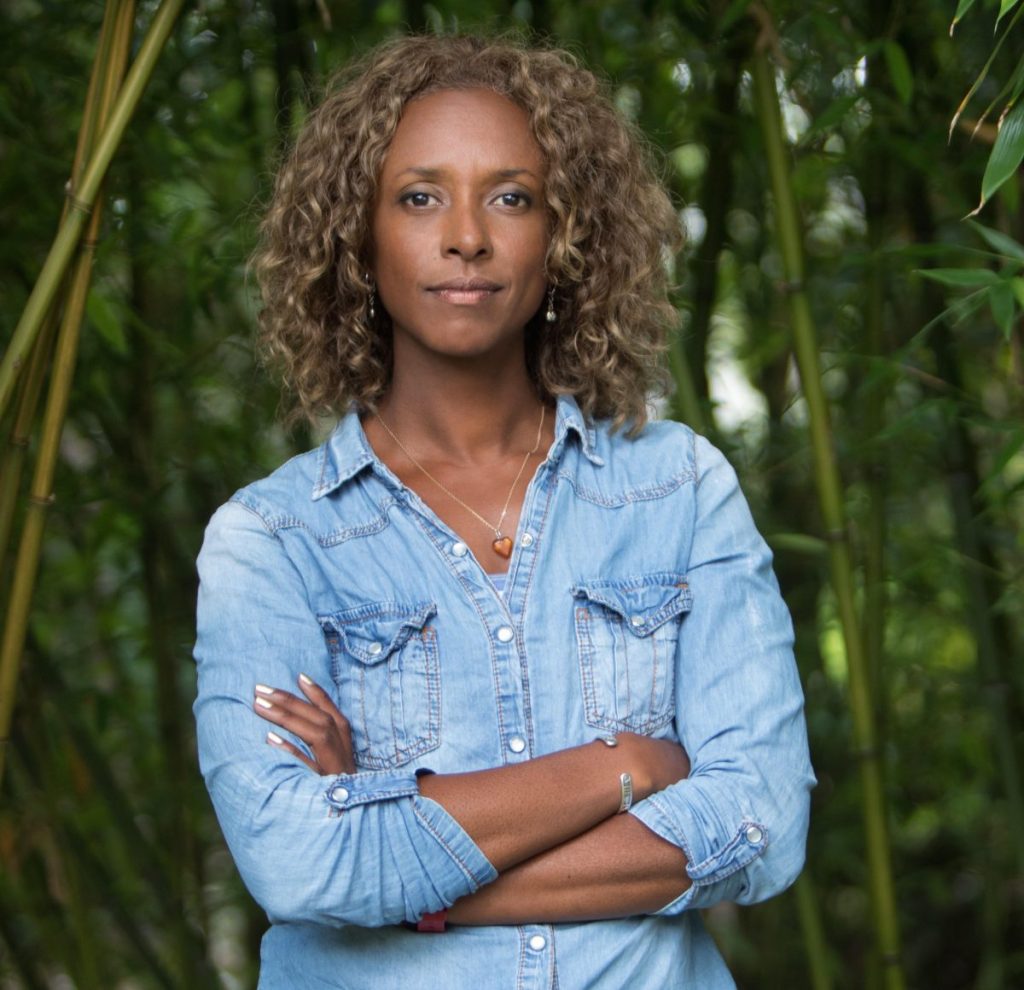 Portrait image of Gillian Burke, she is looking at the camera with her arms folded over her blue denim shirt, a bamboo forest can be seen behind her