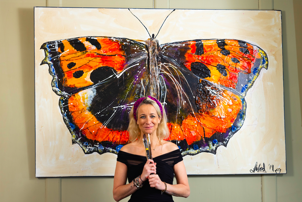 Artist Leah Wood stands in front of her painting of an orange butterfly. She wears a black dress with purple headband and is holding a selection of paintbrushes close to her chest.
