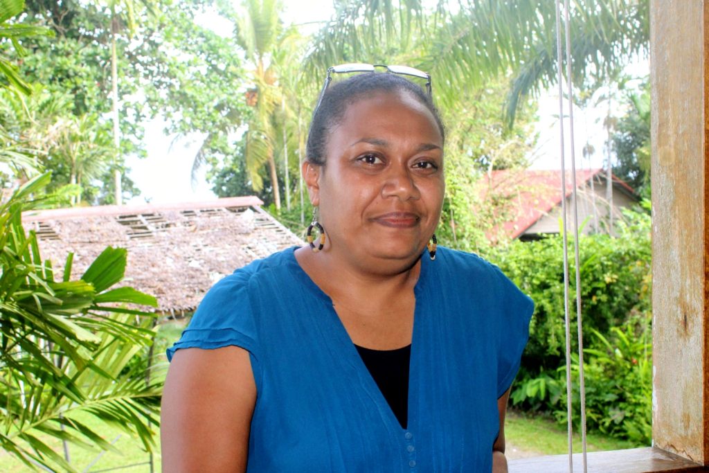 Lelebo poses for her photograph, looking into the camera and smiling, wearing a blue shirt with her glasses perched on top of her head. Behind her are two buildings and many palm trees and other green tropical plants. 