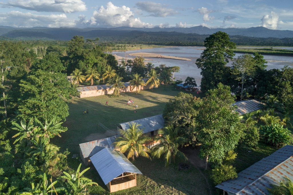 Arial image of Cuitivereni, Peru. Long wooden buildings with metal roofs stand around a large grass pitch, they are surrounded by forest on three sides, while a wide river meets the far edge of the grass pitch. The rainforest can be seen stretching out into the distance.