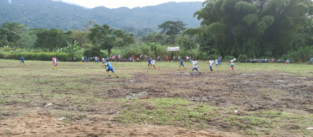 A portrait shot of 13 football plays in the midst of a game. The grassy pitch is muddy in patches, at the edges of the pitch a small audience stands to watch. The pitch is surrounded by lush green forest.