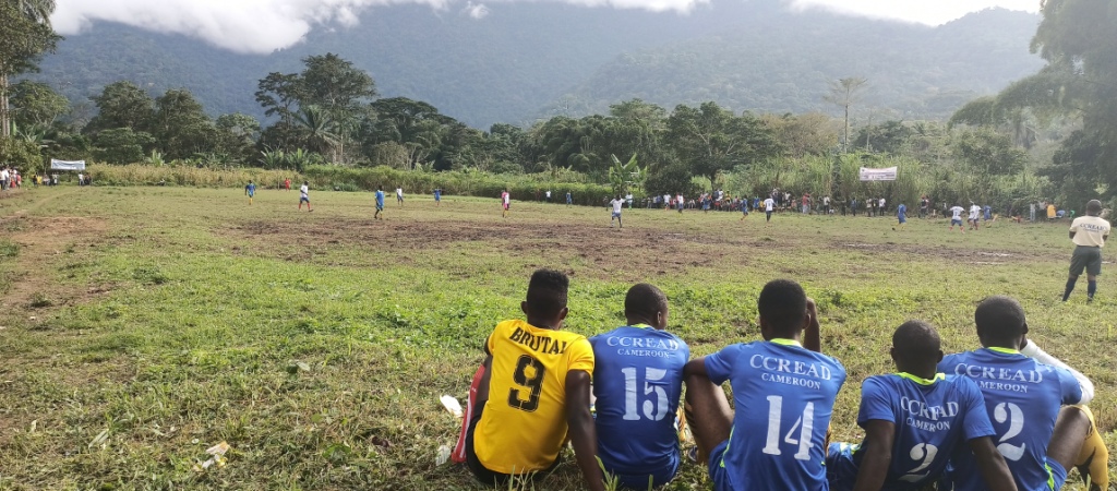 Five football players sit on the side lines of a grassy football pitch, they look onto the pitch watching other players are in the midst of a football game. Green forest surrounds the pitch and continues into the distance.