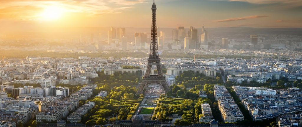 An aerial view of the Eiffel Tower at sunset. Many trees are surrounding the base of. the tower and the city sprawls out behind. 