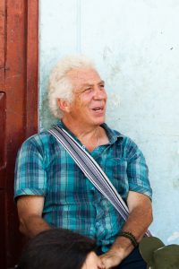 A candid photo of a man looking thoughtfully off camera, wearing a checked shirt and leaning on a pale blue wall.