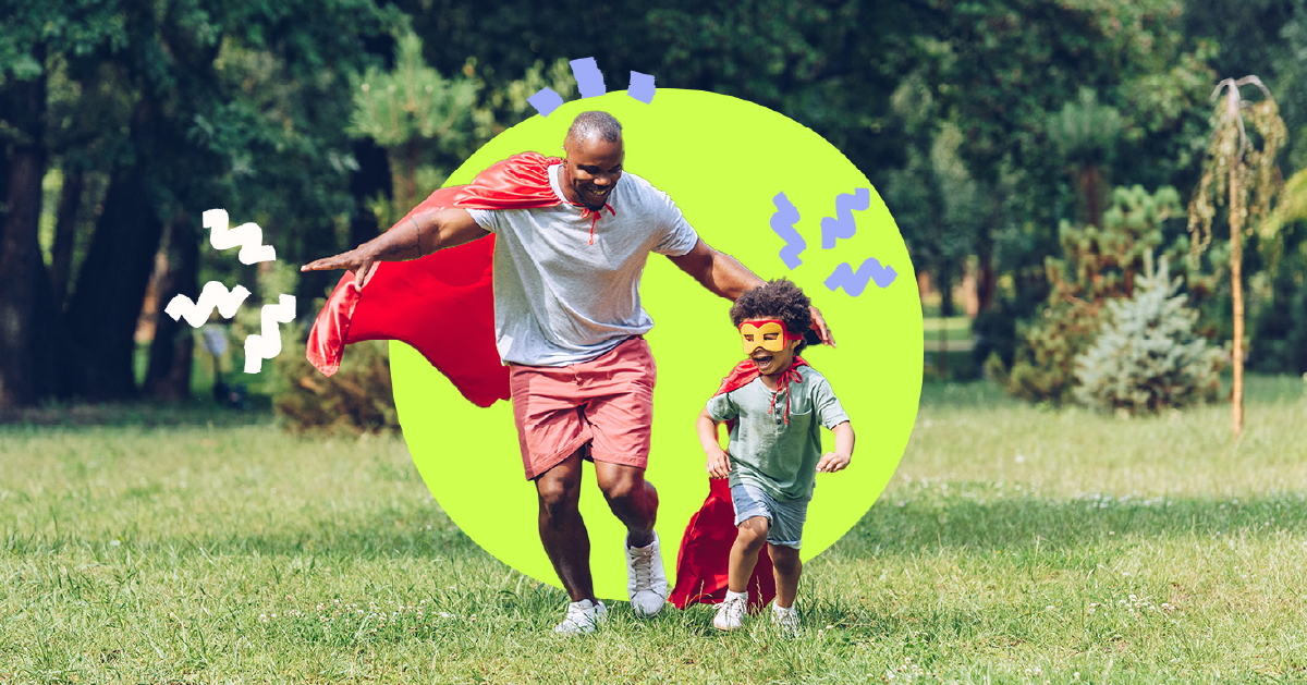 Father and son fun across a grassy field with trees in the back ground. They are both wearing shorts and teeshirts and have bright red superhero capes tied around their necks, the son also sports a yellow superhero mask. 