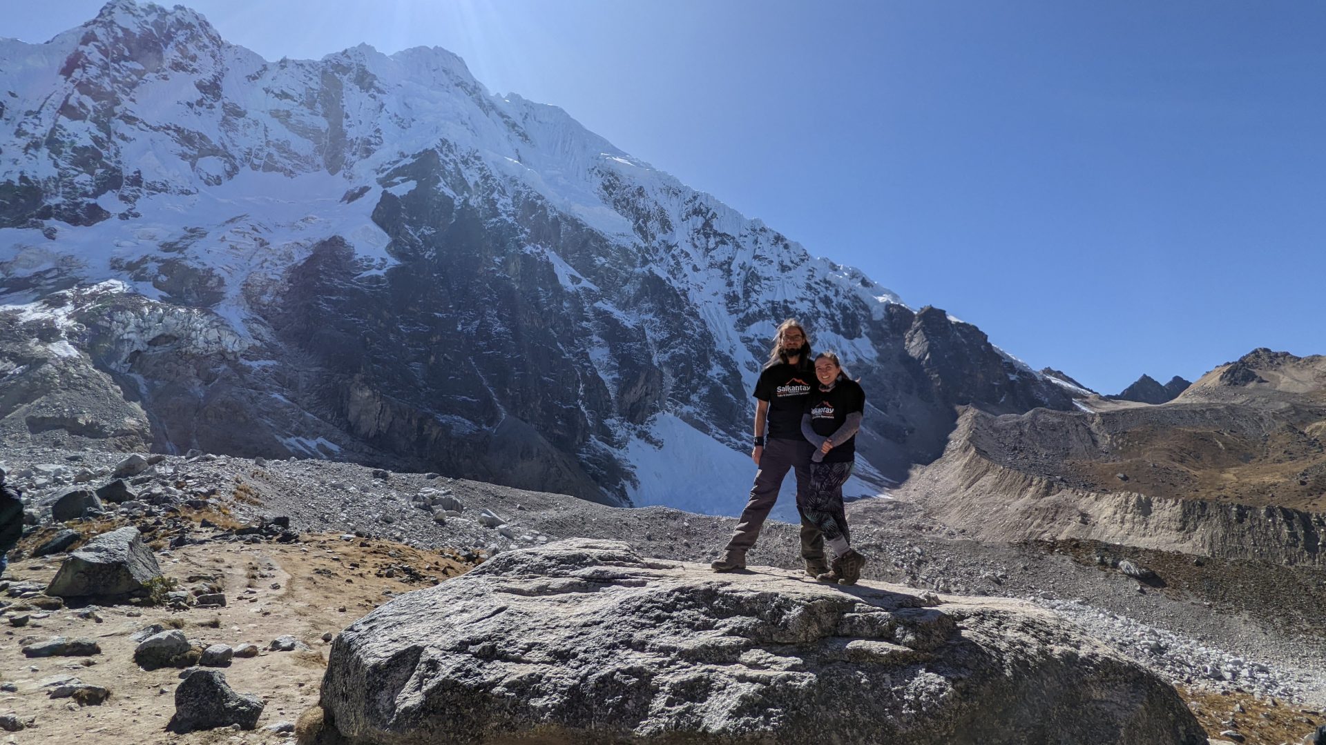 Luke Howkins and his wife smile at the camera and stand together on top of a large boulder in front of a mountain capped in snow. The sun shines above in the cloudless blue sky.
