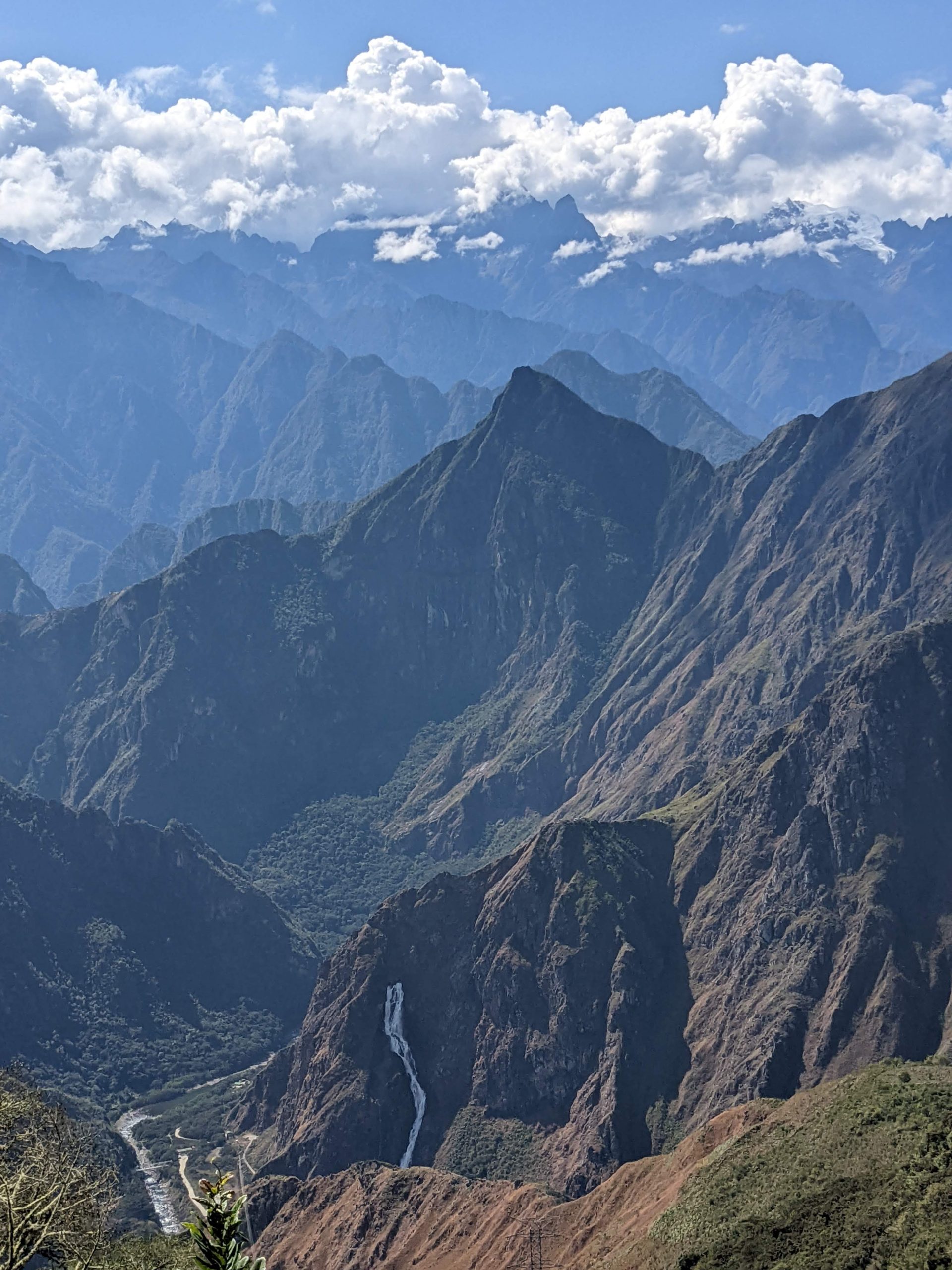 A photo of mountains taken by Luke Howkins. Clouds hang over the tip of the mountain range in the distance. In the forefront a waterfall can be seen bursting from the mountain side.