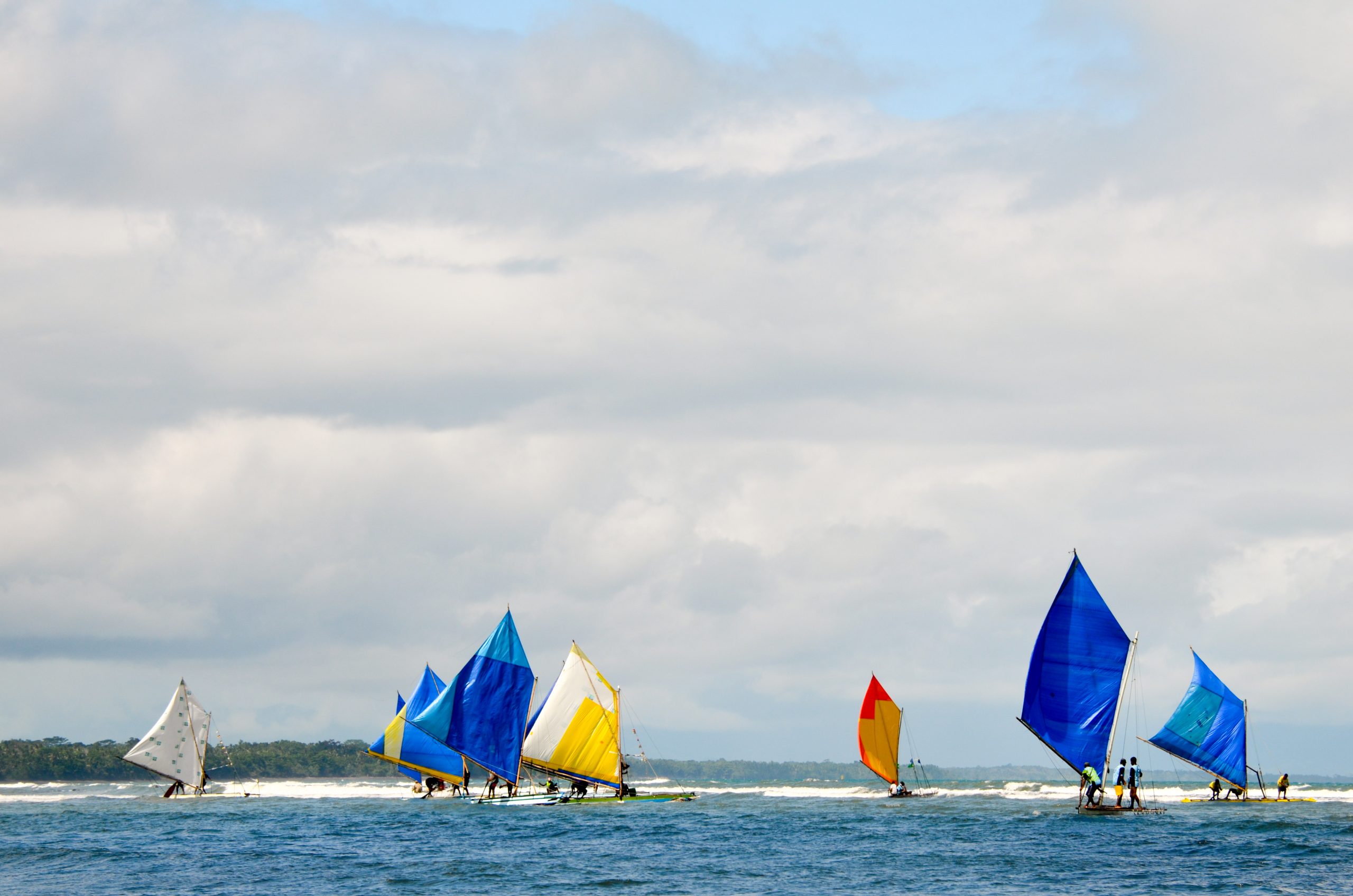 In Gadaisu, Papua New Guinea (PNG), brightly coloured, blue, orange, yellow and white sail boats race out over a blue sea towards a reef break. White clouds hang overhead and rainforest can be seen in the distance.