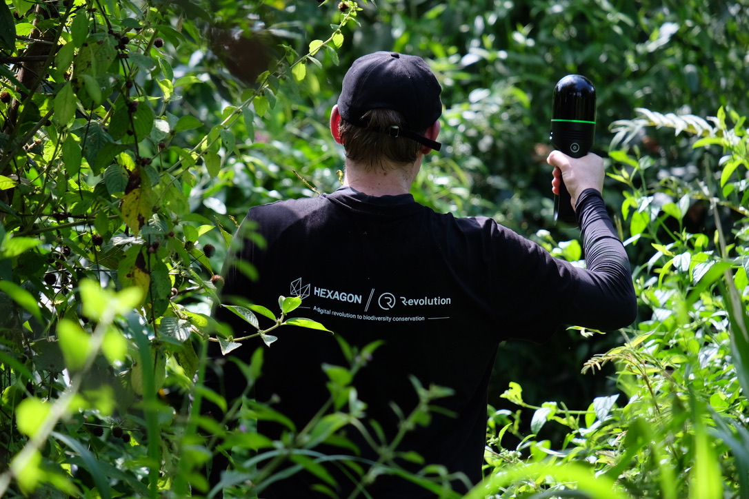 The image shows a person wearing a black top with the "Hexagon" and "Revolution" logos, working in a lush green outdoor environment, collecting forest data using a handheld device while surrounded by dense rainforest vegetation.