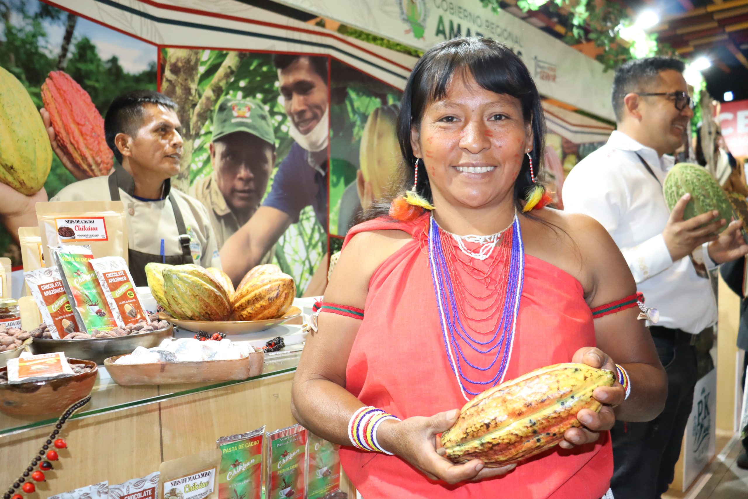 Cemilia Pijushkun part of the post-harvest team smiles at the camera, she is wearing traditional Awajún dress including a red dress, with traditional jewellery. She holds a cacao pod in her hands. Behind her we can see the cacao fair taking place, the Chikáun stall is directly behind her where we can see more cacao pods and the cacao products for sale. 