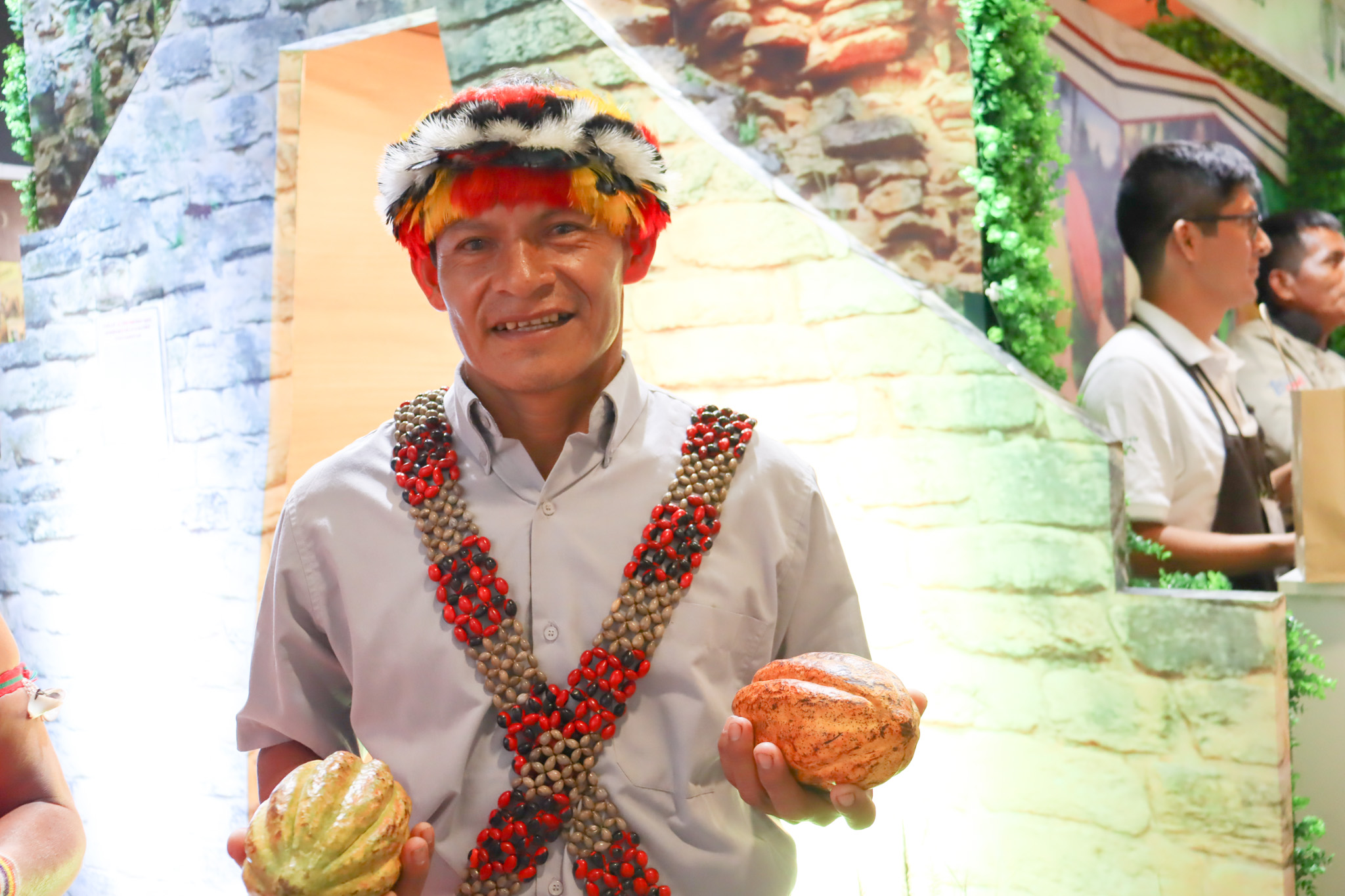 Javier president of Pamuk Bakau association of cacao producers smiles at the camera, he is wearing a traditional dress head pice and holds a cacao pod in each hand. 