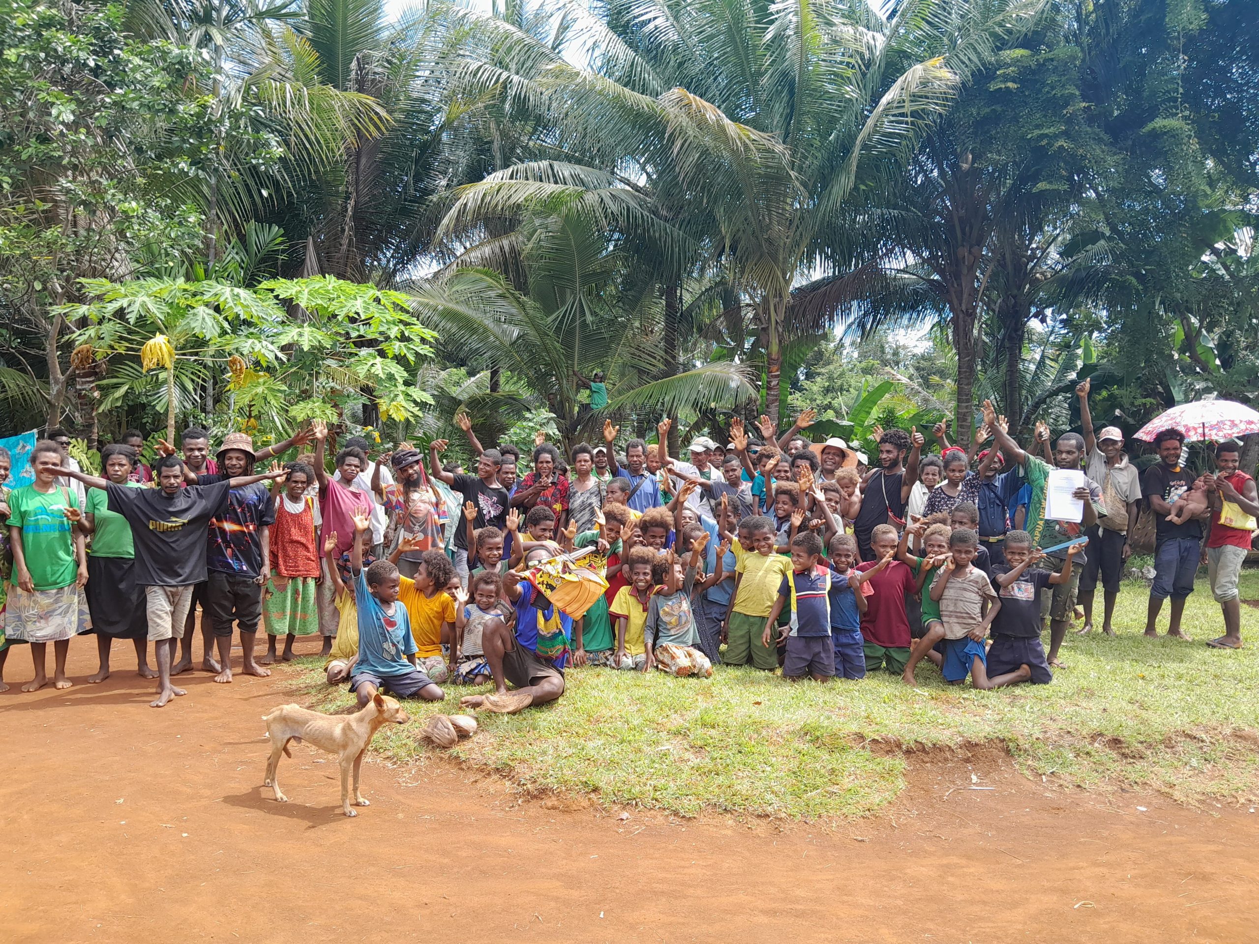 A group of people in Waduada community in Papua New Guinea standnig in front of palm trees and waving their hands 